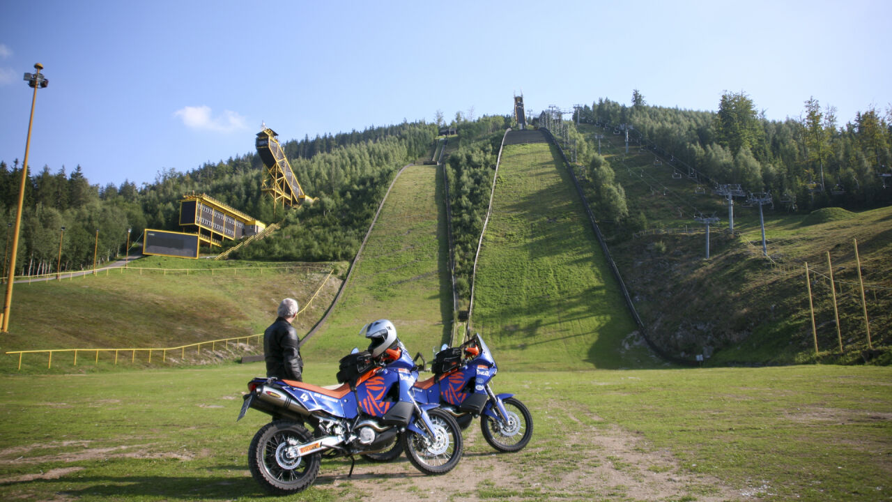 Zwei Reiseenduros vor einer großen Skisprungschanze am Waldhang Zwei Reiseenduros vor einer großen Skisprungschanze am Waldhang