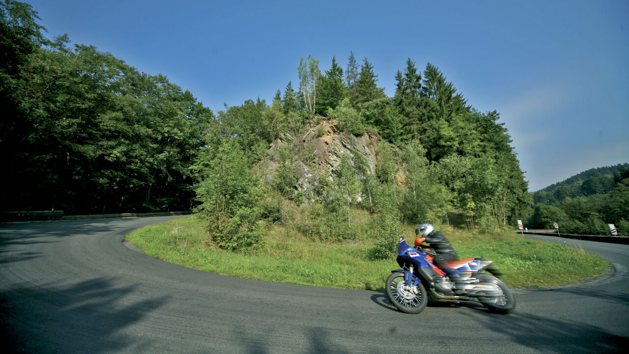 Reiseenduro in einer weiten Rechtskurve vor bewaldetem Hang Reiseenduro in einer weiten Rechtskurve vor bewaldetem Hang